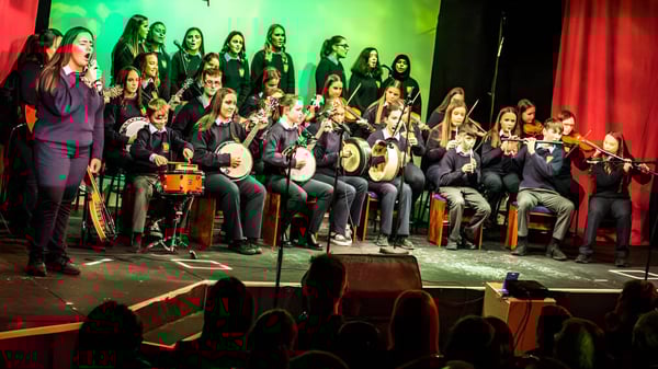 El coro de la Cnoc Mhuire Secondary School actuando en un escenario con iluminación colorida y una cortina roja.