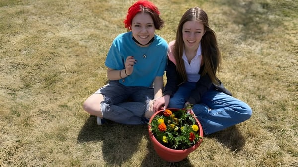 Dos estudiantes de Coalhurst High School están sentadas en el césped y sostienen una colorida cesta con plantas.