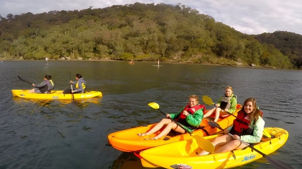 Los estudiantes de la Coláiste an Chreagáin reman en kayaks amarillos en un lago frente a colinas boscosas.
