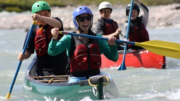 Alumnos de la Coláiste Bhaile Chláir reman con cascos coloridos en una pequeña embarcación verde por un río a través de un paisaje cubierto de hierba.