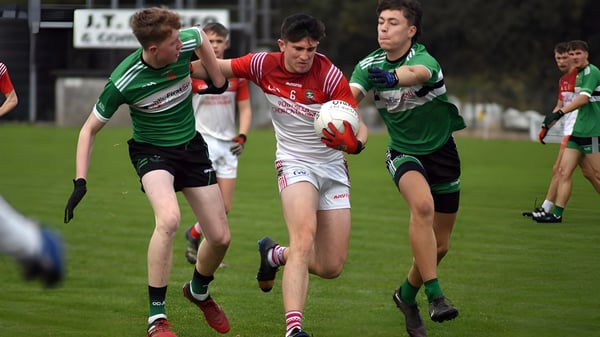 Estudiantes de Coláiste Chríost Rí juegan un partido de fútbol en un campo deportivo con espectadores al fondo.