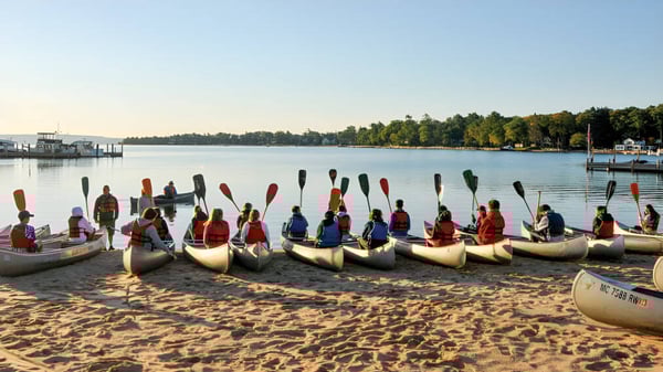 Estudiantes de la Coláiste Éamann Rís reman con canoas en ropa tradicional en un lago tranquilo rodeado de bosque.