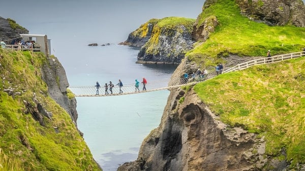 Estudiantes de Coláiste Mhuire (Ballygar) cruzan un puente colgante sobre un paisaje costero con acantilados y agua.