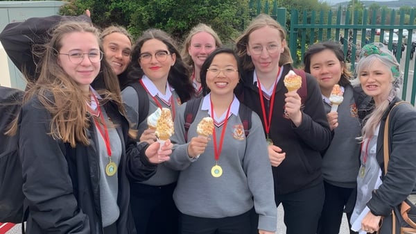 Estudiantes de la Colaiste Muire (Cobh) en ropa de graduación se reúnen con diplomas frente a una cerca verde.