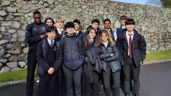 Un grupo de estudiantes en uniformes escolares está de pie frente a una pared de piedra en el terreno de la Colaiste Muire Mathair.
