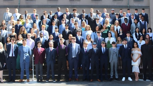 Un grupo de personas vestidas de manera formal está de pie juntas al aire libre frente a un edificio en el campus de la Colaiste Muire Mathair.