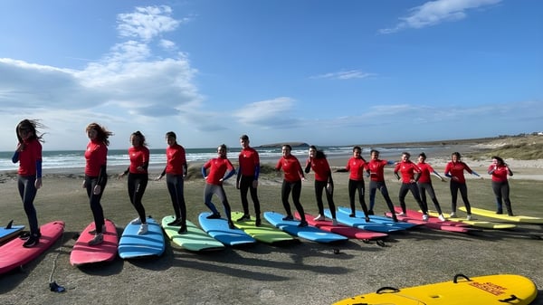 Estudiantes de Coláiste Pobail Acla están en trajes de neopreno rojos en la playa junto a coloridas tablas de surf con un paisaje costero de fondo.