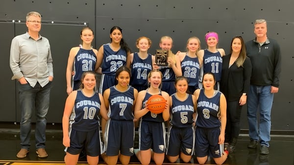 Un grupo de jóvenes jugadoras de baloncesto en uniforme se encuentra juntas en el gimnasio de la College Heights Secondary School.