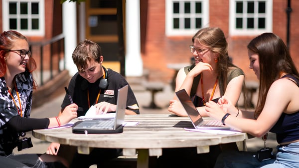 Estudiantes del College of Richard Collyer discuten al aire libre en una mesa de madera con laptops.