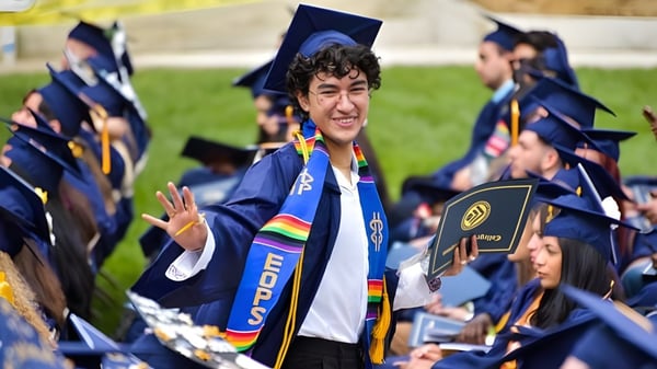 Un graduado en toga y birrete azules está con otros graduados en el campus del College of the Canyons al aire libre.
