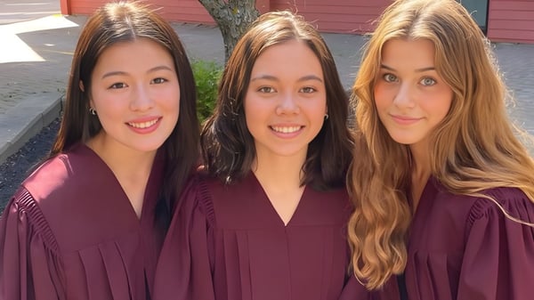 Tres graduadas en togas posan frente a un edificio de madera roja en el campus de la Colonel Gray High School.