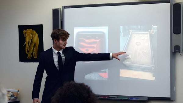 Un hombre en traje está frente a una gran pantalla y señala una proyección en el campus de la Colorado Rocky Mountain School.