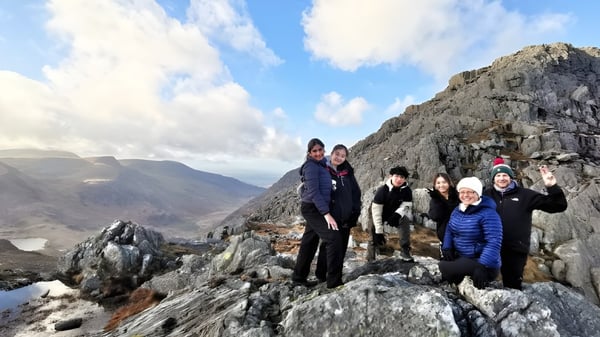 Un grupo de estudiantes del Concord College está en un saliente rocoso con montañas y un cielo nublado al fondo.