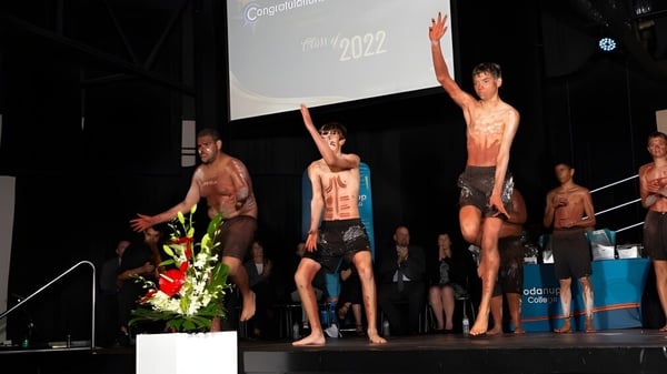 Un grupo de jóvenes sin camisa está en el escenario en el campus del Coodanup College con una gran pantalla de fondo que muestra el año 2022.