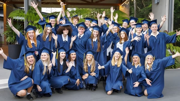 Un gran grupo de graduados celebra con togas y birretes azules en el campus de la Coolum State High School.