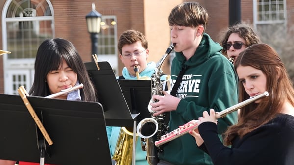 Estudiantes de la Corfe Hills School tocan al aire libre varios instrumentos musicales y leen partituras frente a un edificio de ladrillo.