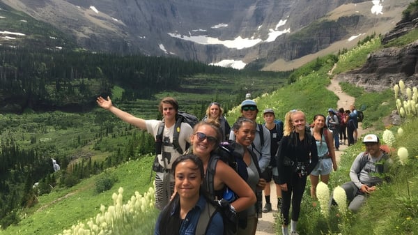 Estudiantes de la Corfe Hills School caminan por un sendero en un paisaje montañoso con vegetación verde y rocas al fondo.