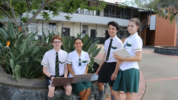 Cuatro estudiantes de la Corinda State High School están de pie frente al edificio escolar rodeados de vegetación verde.