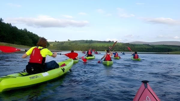 Alumnos de la Cork Educate Together Secondary School reman en coloridos kayaks en un lago tranquilo rodeado de bosques verdes.