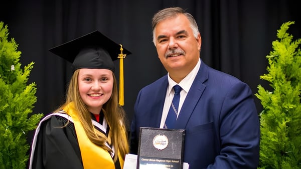 Una graduada está junto a un hombre en traje frente a un fondo verde en el terreno de la Corner Brook Regional High School.