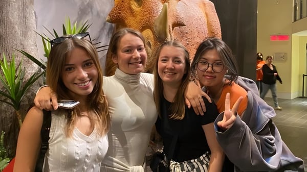 Cuatro estudiantes de la Cornwall Collegiate & Vocational School posan juntas frente a coloridas obras de arte en el interior.