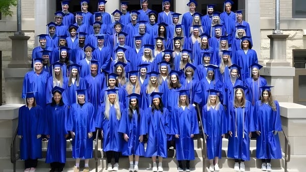 Un gran grupo de graduados de las Cotter Schools está de pie en togas y birretes azules en los escalones de un edificio con fachada de piedra.