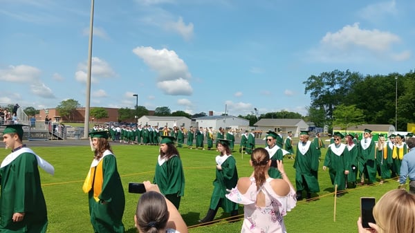 Graduadas y graduados de Coventry High School en togas verdes y amarillas están de pie en un prado frente a un edificio de ladrillo.