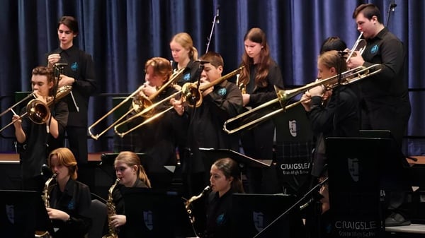 Un grupo de estudiantes de la Craigslea State High School toca en el escenario frente a un telón oscuro con varios instrumentos.
