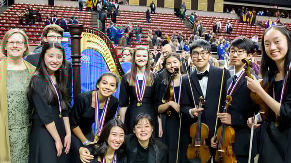 Un grupo de jóvenes músicos posa en el escenario del Cranbrook Education Campus frente a asientos rojos y un gran arpa.