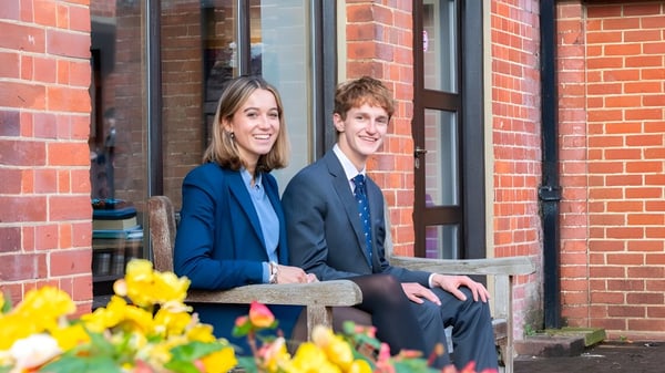 Dos alumnas y alumnos de la Cranleigh School están sentados juntos en un banco frente a un edificio de ladrillo con flores amarillas.