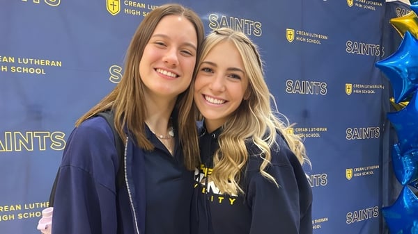 Dos estudiantes sonrientes con cabello largo y rubio están de pie frente a un fondo con el logo de la Crean Lutheran High School.