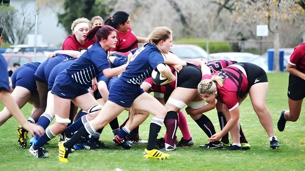 Estudiantes de la Crescent Heights High School están en un scrum de rugby en un campo de césped con árboles y edificios al fondo.