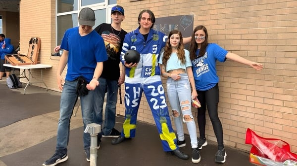Un grupo de alumnas y alumnos de la Crescent Heights High School está de pie en camisetas deportivas frente a un edificio de ladrillo.