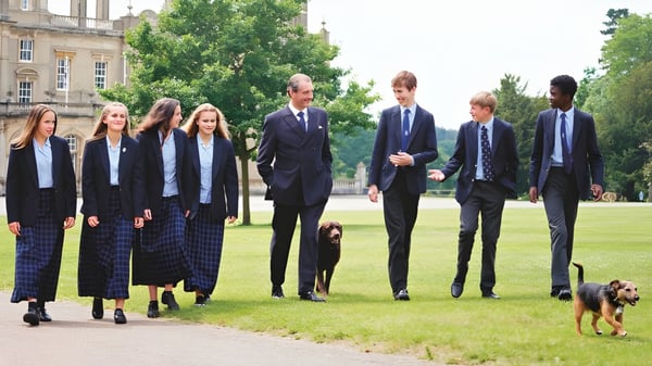 Un grupo de estudiantes de la Culford School camina junto con un perro por una pradera entre árboles y edificios.
