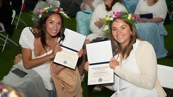 Dos estudiantes de la Dana Hall School llevan vestidos blancos y coronas de flores y sostienen sus diplomas en una celebración al aire libre.