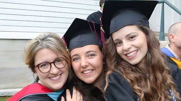 Tres jóvenes mujeres en vestimenta de graduación de la Darrow School están frente a un edificio de madera.