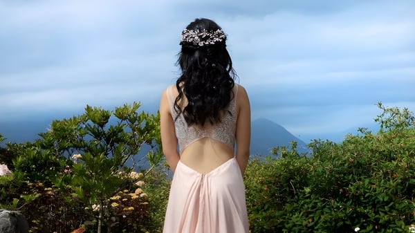 Persona con cabello largo y oscuro en vestido rosa se encuentra en un paisaje verde frente a montañas en el terreno de la David Thompson Secondary School.