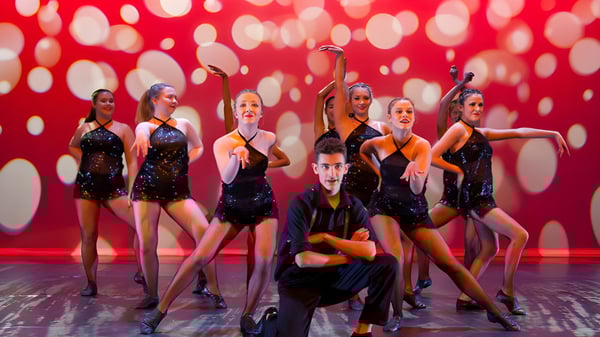 Un grupo de bailarinas y bailarines de la Davidson High School se presenta en un escenario frente a un fondo rojo con puntos blancos.