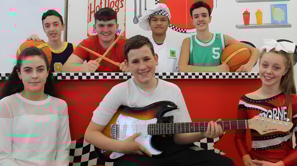 Un grupo de estudiantes con instrumentos musicales y equipos deportivos frente a un colorido patrón de cuadros en el campus del Davitt College.