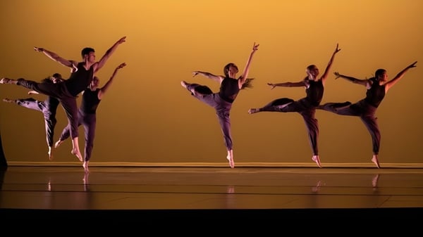 Un grupo de bailarinas y bailarines de la Deerfield Academy realiza una actuación sincronizada en el escenario frente a un fondo dorado.