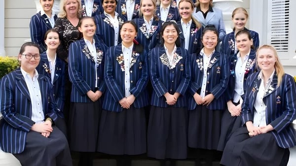 Un grupo de alumnas de la Diocesan School For Girls está en ropa de graduación frente a un edificio con columnas blancas.