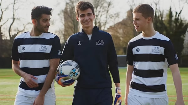 Tres estudiantes del DLD College London están juntos en un campo de césped con árboles y un cielo nublado de fondo.