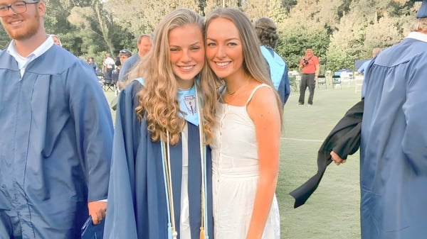 Dos estudiantes de la Dominion Christian School están de pie en toga de graduación sobre un prado.