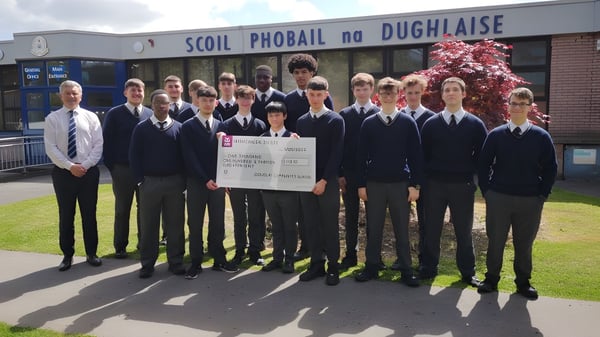 Estudiantes de la Douglas Community School están en uniformes escolares frente al edificio escolar sosteniendo un gran cheque.