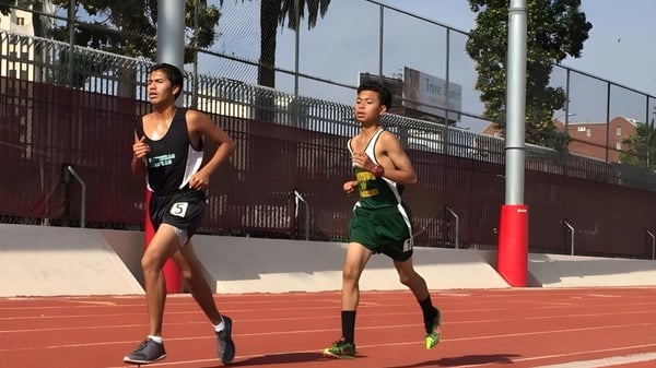 Dos atletas de la Downtown Magnet High School corren en la pista deportiva roja con una cerca y edificios de fondo.