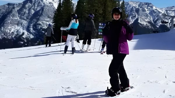 Una estudiante de la Dr. Elliott School lleva equipo de esquí en una pista nevada con montañas y pinos de fondo.