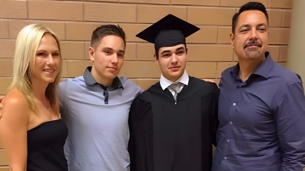 Un grupo de cuatro personas, incluyendo a un estudiante en una toga de graduación, está frente a una pared de ladrillos en el terreno de la Dr. Frank J. Hayden Secondary School.