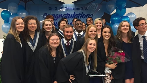 Estudiantes de la Dr. G.W. Williams Secondary School se reúnen en togas de graduación frente a un banner de felicitación con un paraguas azul encima.