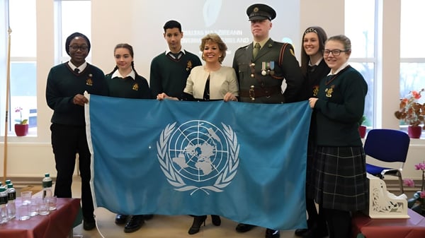 Estudiantes de la Drogheda Grammar School y personas en uniformes militares sostienen juntos una bandera de las Naciones Unidas en un interior.