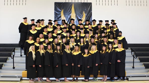 Un gran grupo de estudiantes de la Drumheller Valley School lleva vestimenta de graduación negra frente a pancartas amarillo-azules.
